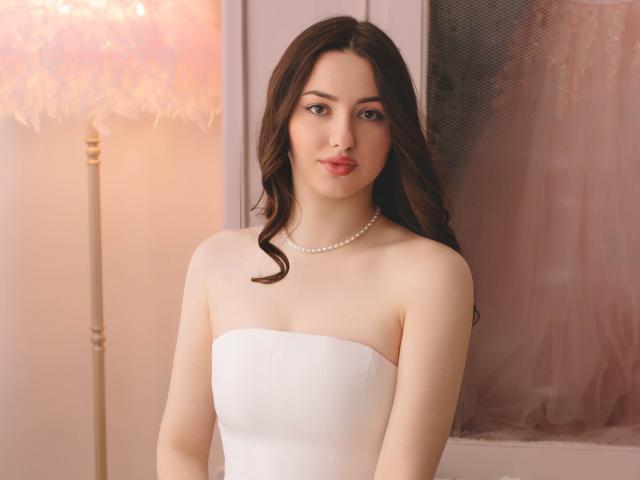 Photographie de ChristinaBliss avec de longs cheveux noirs ondulés, portant une robe blanche sans bretelles et un collier de perles, debout dans devant un arrière-plan légèrement éclairé, aux tons chauds. Tension sexuelle, regard séduisant, attirant..