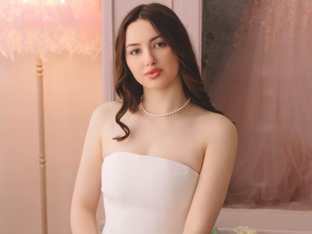 Foto van ChristinaBliss met lang, donkerbruin haar, gekleed in een witte jurk zonder bandjes en een parelketting. Zacht licht van een lamp aan de linkerkant, subtiele roze gordijnen op de achtergrond. Absoluut prachtig.