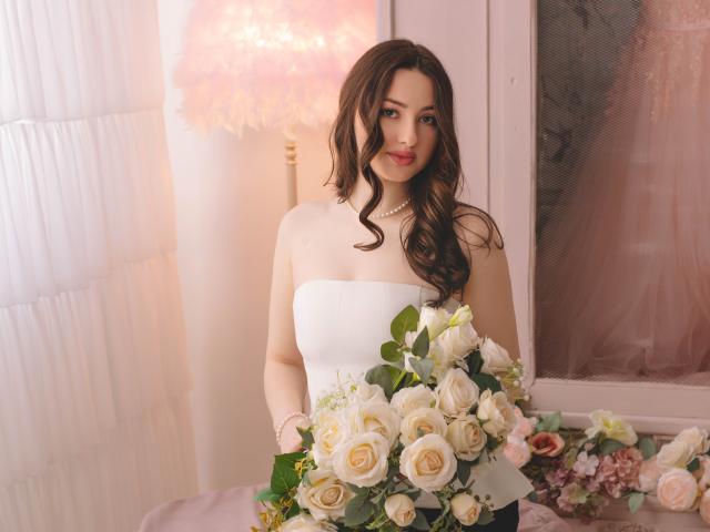 Fotografia de ChristinaBliss, em pé em um quarto com iluminação suave, segurando um grande buquê de rosas brancas e rosa. Ela tem cabelos longos, cacheados e pretos, lábios vermelhos e um vestido branco sem alças..