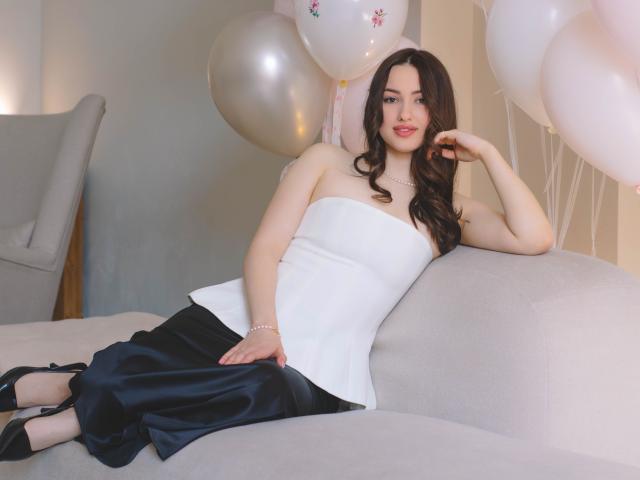 ChristinaBliss ligt ontspannen op een grijze bank, gekleed in een wit topje zonder bandjes en een zwarte rok, omringd door witte ballonnen. Ze heeft lang, golvend zwart haar en een zelfverzekerde, aantrekkelijke uitdrukking..