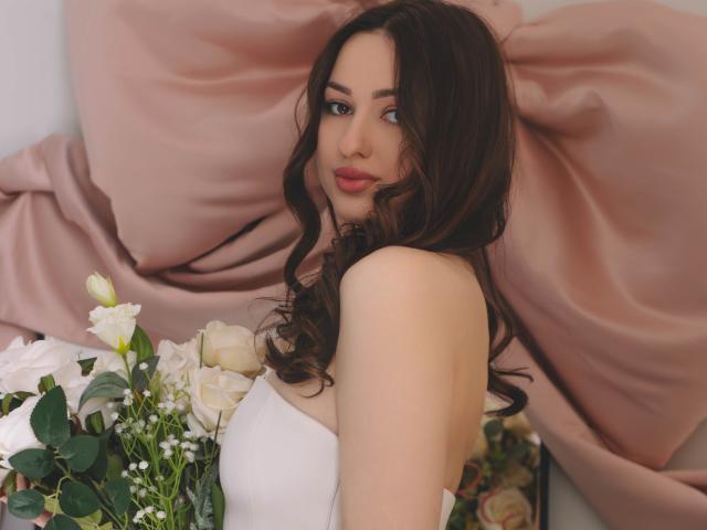 Fotografía de ChristinaBliss, una mujer morena con cabello ondulado, vestida con un vestido blanco sin tirantes, sosteniendo un ramo de flores blancas y amarillas. flores, fondo de tela rosa drapeada. Mirada sensual y directa, ligera sonrisa, iluminación suave..