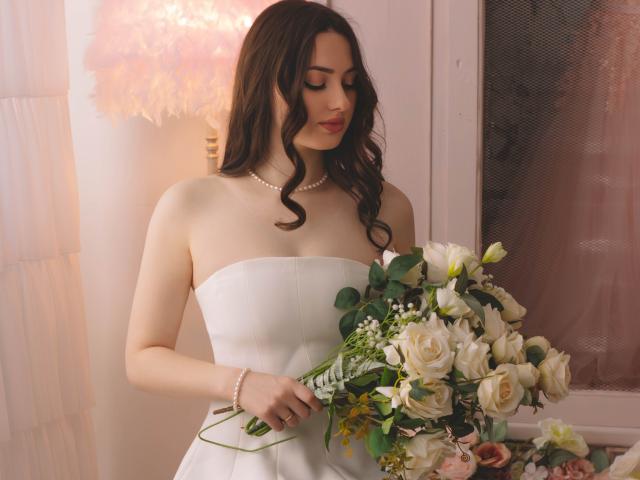 Fotografia di ChristinaBliss con lunghi capelli scuri, che indossa un abito da sposa bianco senza spalline e una collana di perle, mentre tiene in mano un... Un bouquet di rose bianche e rosa. Lei guarda in basso con un sorriso appena accennato.