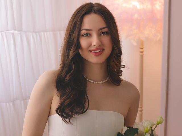 Fotografía de ChristinaBliss con cabello castaño largo y ondulado, vestida con un vestido blanco sin tirantes y un collar de perlas. Ella está sonriendo, con flores blancas en el primer plano a la derecha. Iluminación suave y cálida. Hermosamente increíble..