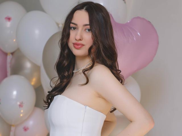 Foto van ChristinaBliss, een aantrekkelijke vrouw met lang, golvend zwart haar, die een witte jurk zonder bandjes en een parelketting draagt. ketting.
ze staat met haar hand op haar heup, omringd door witte en roze ballonnen. Heel aantrekkelijk, met een zelfverzekerde, verleidelijke blik..