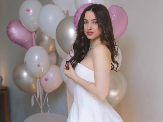 Fotografia di ChristinaBliss, in piedi con il profilo laterale, che indossa un abito bianco senza spalline, tenendosi il seno. Circondato da palloncini rosa e bianchi. I suoi lunghi capelli neri, il rossetto rosso e la collana di perle..