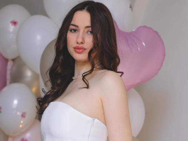 Photograph of ChristinaBliss, a fair-skinned woman with long, wavy black hair, wearing a strapless white dress, standing in front of pink and white balloons. She has a sultry expression.