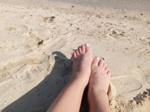 Nahaufnahme von GladisBelles nackten Füßen, die auf einem sandigen Strand stehen, wobei Schatten auf den Boden fallen. Die Füße leicht auseinander, Sand zwischen den Zehen. Helles Sonnenlicht, feine Textur der Sandkörner..