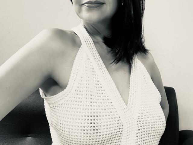 Black and white photo of LorenHolmes with shoulder-length dark hair, wearing a white, knitted halter top, showcasing her medium-sized breasts. LorenHolmes has a slight, seductive smile. High contrast.