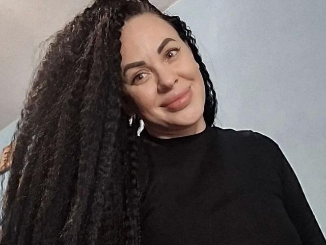 Fotografía de Eleonoracherry-hot, sonriendo con una ligera inclinación, cabello negro y rizado largo, vistiendo una camisa negra, piel clara. ojos marrones, maquillaje sutil, luz suave, fondo ligeramente desenfocado. Qué lindo/a..