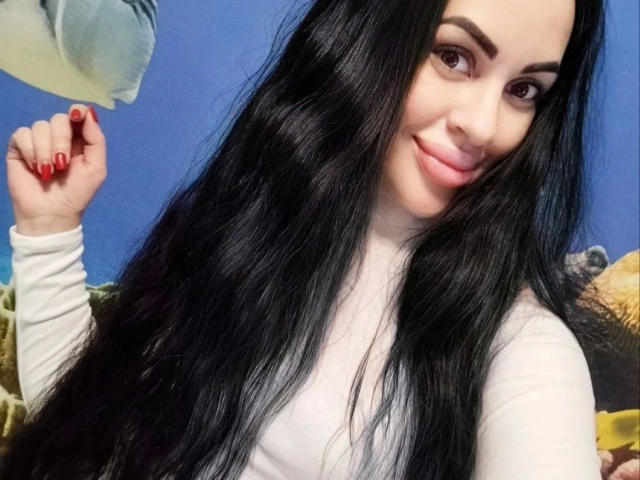 Close-up selfie of Eleonoracherry-hot with long, black hair, white top, red nails, and a playful smirk. Blue background, raised hand with red nail polish. Sexual tension, flirty vibe.