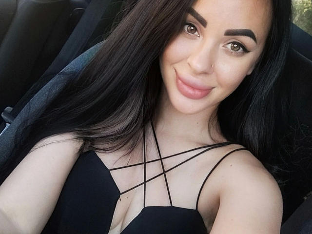 Fotografia ravvicinata di Eleonora, con capelli neri lunghi, occhi marroni e un piccolo piercing al naso. Indossa una canottiera nera con spalline sottili, sorride leggermente ed è seduta in un'auto. Rapporti con una sensualità accattivante..