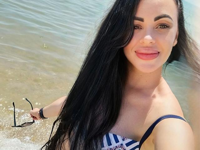 Fotografia di Eleonoracherry-hot, che sorride alla macchina fotografica con lunghi capelli neri, indossa un bikini a righe bianche e nere. in cima, in piedi in acque poco profonde. Ha un aspetto seducente e attraente..