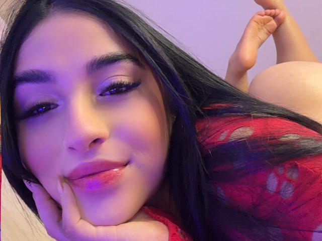 Primer plano de EmmaWaltter acostada boca abajo, con la cabeza apoyada en la mano, cabello negro, blusa roja con estampado floral, y el trasero desnudo levantado. el fondo, con una sonrisa burlona, iluminación morada-azul, tensión sexual.