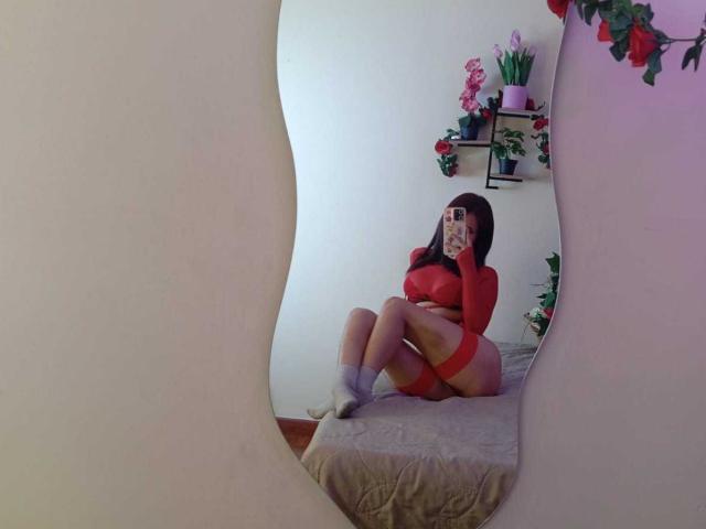 KatheLux, sitting on a beige bed, wearing a red long-sleeve top and red shorts, white socks, legs crossed, holding a phone, room with pink flowers, mirror framing the scene.