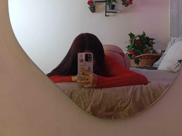 KatheLux, long brunette hair, red top, lying face down on beige bed, holding phone, mirror reflection, potted plants, flowers on wall, casual, sexual tension, phone partially obscures face.