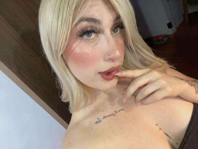 Close-up of SammyViolet, a blonde woman with heavy makeup, including long eyelashes and pink blush. She's biting her finger, showing off tattoos on her chest. Her expression is seductive.