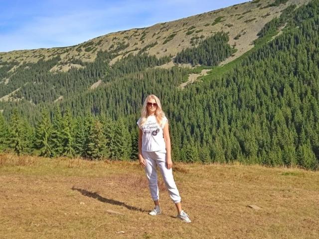 GladisBelle stands on a grassy hill, wearing white pants and a tank top, with sunglasses, against a backdrop of dense green trees and rolling mountains under a bright blue sky.