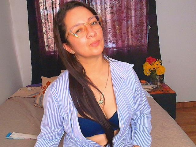 VictoriaRousse with long black hair, wearing glasses and a blue-striped shirt over a black bra, sitting on a bed. Background: dark curtains, bedside table with yellow flowers. Sexual tension: subtle, inviting.