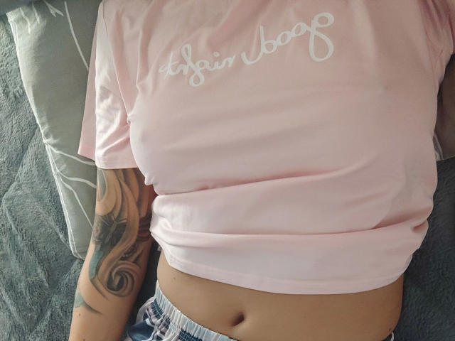 Gros plan de Svenjah1-hot allongée sur un canapé, portant un t-shirt rose avec l'inscription "stay boo" en blanc, en lettres cursives. Tatouage visible sur le bras droit, ventre apparent, pantalon gris. Tension sexuelle.