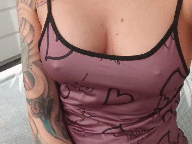 Primo piano del petto e della parte superiore del corpo di Svenjah1-hot, che indossa una canotta trasparente color malva con motivi a cuore e scritte nere. Contorno del capezzolo visibile attraverso il tessuto. Tatuaggio dettagliato sul braccio destro. Tensione sessuale.