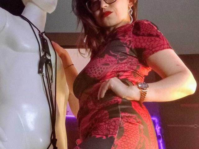 AngelinaBlanche in a tight red lace dress, leaning forward with hand on hip, showing off her curves. She's wearing a watch and earrings, with a mannequin in the foreground. Sexual tension.