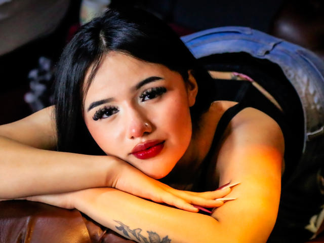 Fotografia ravvicinata di MiaTanya con i capelli scuri, sdraiata su un divano in pelle marrone, con le braccia incrociate sul petto, labbra rosse, tatuaggio. sulla parte anteriore del braccio, sguardo intenso, pelle luminosa, aspetto seducente..