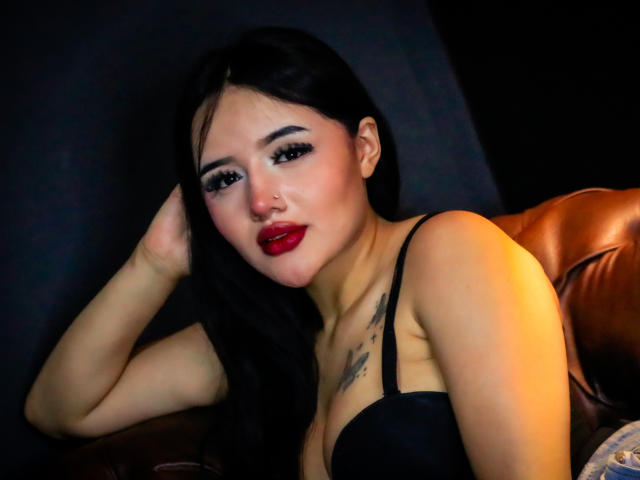 MiaTanya with black hair, red lipstick, and black bra, lying on a brown leather chair, hand in hair, intense gaze, dark background, sexy, sultry expression, visible chest tattoo.