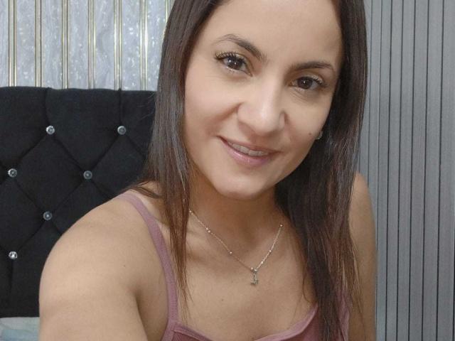 Close-up de SolaraDubois com cabelo castanho liso, pele clara e um leve sorriso. Ela está vestindo uma camiseta rosa e um colar com um pequeno pingente. Cabeceira preta ao fundo. Incrivelmente atraente..