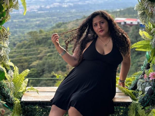 Genesis Montenegro, com cabelos escuros e cacheados, vestindo um vestido preto, sentada em um banco de madeira, segurando o cabelo, em meio a um verde exuberante. folhagem, montanhas ao fundo, sensual, confiante, luz natural, fundo ligeiramente desfocado..