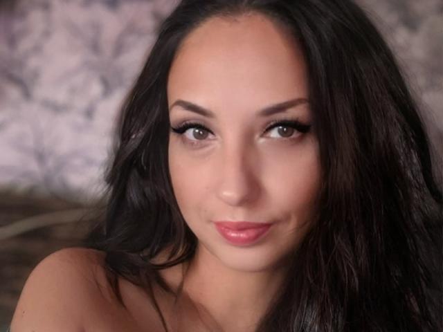 Fotografia ravvicinata di Emiliya Philips, una donna con lunghi capelli neri, sopracciglia scure e rossetto rosso, che guarda direttamente. la fotocamera, con un sorriso appena accennato, una luce soffusa che mette in risalto il suo viso.