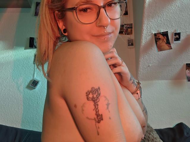 Fotografia di MelliSexy97-hot con capelli biondi, occhiali e un piercing al naso, senza top, che mostra un tatuaggio a forma di chiave sulla spalla. Sta guardando la fotocamera con un sorriso appena accennato, con le mani vicino al collo..