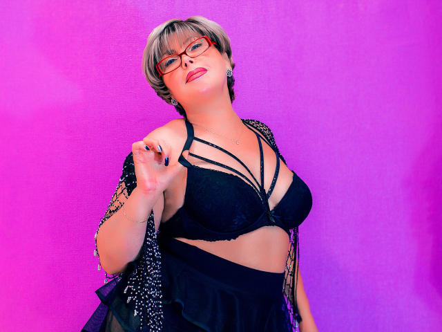 Digital portrait of LonelyMonika, short gray hair, red glasses, black bra with strappy details, black dress, leaning forward with hand near face, neon pink and purple gradient background, sexy pose.
