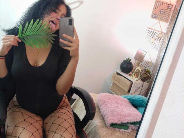 AriaVega, with curly black hair and tan skin, sits on a chair, holding a green leaf over her mouth, wearing a black top, fishnet stockings, and tongue out, taking a mirror selfie.