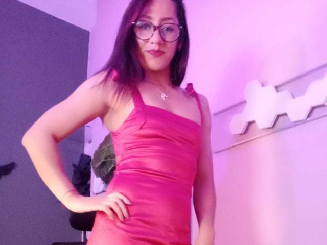 SamanthaCorine, a hot Asian chick with glasses, stands confidently in a tight red dress, hand on hip, wearing a necklace, with purple and white lighting, creating a sexy vibe.