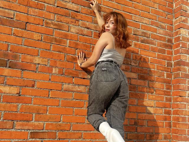 Emily Klein mit lockigem, braunem Haar, einem weißen Tanktop und hoch taillierten, blauen Jeans, lehnt an einer roten Backsteinmauer, rechts. Hand angehoben, linke Hand an der Wand, weiße Socken, sexuelle Spannung..