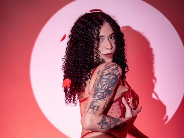 AnnieRoselle with curly black hair, red lingerie, tattoos on right arm, holding her tits, red petals in hair, looking at camera, shadow of her face on pink background.