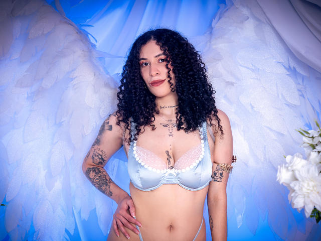 AnnieRoselle with curly black hair, light blue lingerie, and white angel wings, showing tattoos, standing with hands on hips, looking at camera, blue light, white flowers on right.