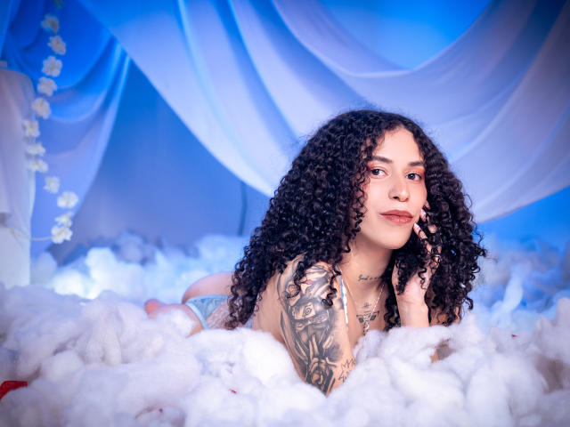 AnnieRoselle with curly black hair, tattoos on her right arm, lying in fluffy white clouds, wearing blue panties, looking at the camera, touching her cheek with her left hand.