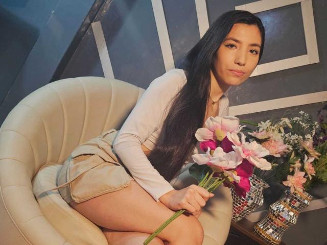 MiaLovelyn, eine asiatische Frau mit langen, schwarzen Haaren, sitzt auf einem beigen Stuhl, trägt ein weißes Oberteil und beigefarbene Shorts und hält etwas Rosa. und weiße Lilien, dunkler Hintergrund, sexuelle Spannung, Blumenvase.