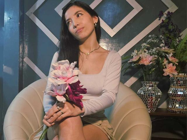 MiaLovelyn with long black hair, sitting on a beige chair, holding a bouquet of pink and white carnations. She's wearing a tight white top and beige shorts. Background includes diamond-patterned wall and floral vases. Fucking sexy, looking up with a slight smile.