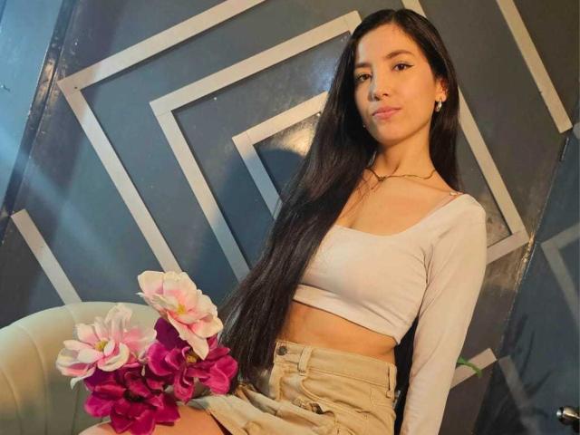 MiaLovelyn, une femme asiatique mince avec de longs cheveux noirs, porte un débardeur blanc et un short beige, et tient un objet rose et rouge. fleurs. Fond avec un motif géométrique. Pose incroyablement séduisante et pleine d'assurance..