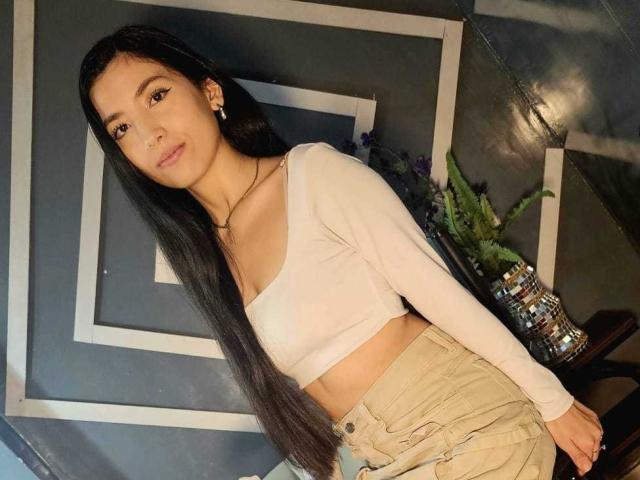 MiaLovelyn stands in front of a geometric wall, leaning slightly forward with long black hair, wearing a white crop top and beige pants. Her expression is neutral, with a slight smile.