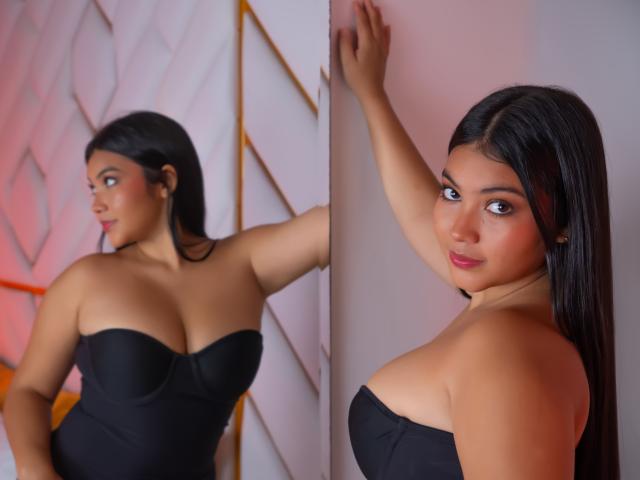 Zwei Fotos von AlisBella mit langen, schwarzen Haaren, die ein schwarzes Kleid ohne Träger trägt und ihre großen Brüste zur Schau stellt. Sie lehnt sich mit einer Hand an eine Wand, die andere Hand oben, und blickt verführerisch in die Kamera. Helle Beleuchtung, roter und weißer Hintergrund.