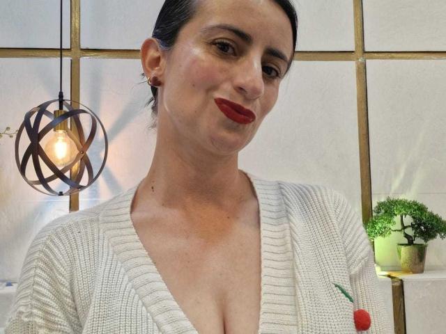 Photographie de BrendaCantorel, une femme aux cheveux foncés, avec du rouge à lèvres, portant un pull blanc texturé à col en V, laissant légèrement entrevoir sa poitrine. Lumière géométrique suspendue, plante verte sur une étagère, pose décontractée et confiante, tension sensuelle, léger sourire..