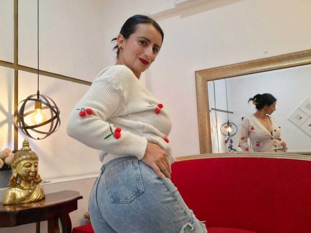 BrendaCantorel, em pé com uma mão no quadril, vestindo um suéter branco com estampas de rosas vermelhas e jeans rasgados, refletido em um espelho. Buda dourado e luminária moderna ao fundo. Incrivelmente sexy..