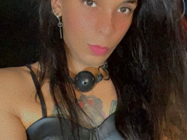 Primo piano di MiissValent con lunghi capelli neri, che indossa una canottiera in pelle nera, un collare nero con un "ball gag" e orecchini. Tatuaggio sul petto. Espressione seria, leggera tensione sessuale..