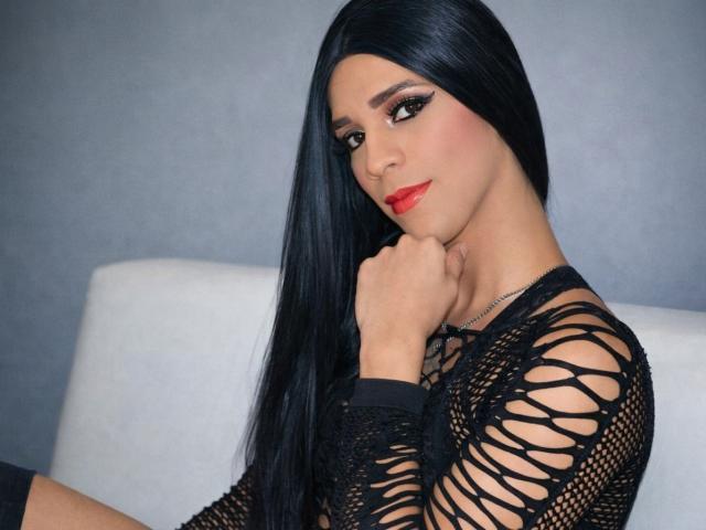 Fotografía de MiissValent con cabello largo y negro, vistiendo una blusa de malla negra, lápiz labial rojo y un maquillaje de ojos llamativo, en posición relajada. con el mentón apoyado en la mano, mirando de forma seductora a la cámara.