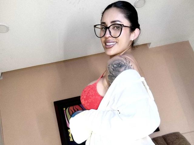 Photographie d'Angelica, cheveux foncés attachés en queue de cheval, portant des lunettes noires et un soutien-gorge en dentelle rouge, avec un tatouage visible. Le bras levé et souriante, avec une chemise blanche qui la couvre partiellement. Extrêmement séduisant..