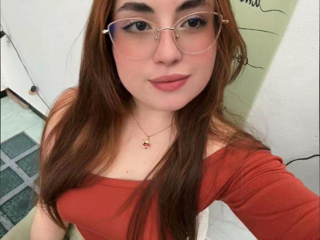 VicluX with long brown hair, wearing a red off-shoulder top and clear glasses, looking directly at the camera. She has a small pendant necklace, a slight smirk, and visible cleavage.