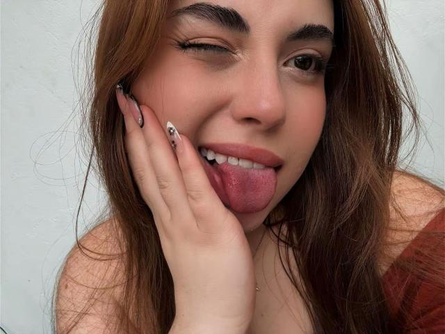 Photo en gros plan de VicluX, avec des cheveux bruns, qui cligne de l'œil, tire la langue et touche sa joue avec une main manucurée. VicluX a la peau claire, des sourcils foncés, et porte un haut rouge. Incroyablement sexy..